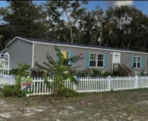 A gray mobile home with blue shutters and ferns around it