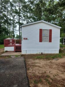 The outside, side of a white mobile home with red shutters and red wooden steps to the entrance, with small paves driveway.