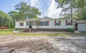 Large white mobile home with brick colored shutters, brick bottom, and brick stairs to entryway with cement sidewalk and open grass in the front yard.