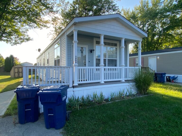 A gray singlewide mobile home with 2 blue trash bins out front
